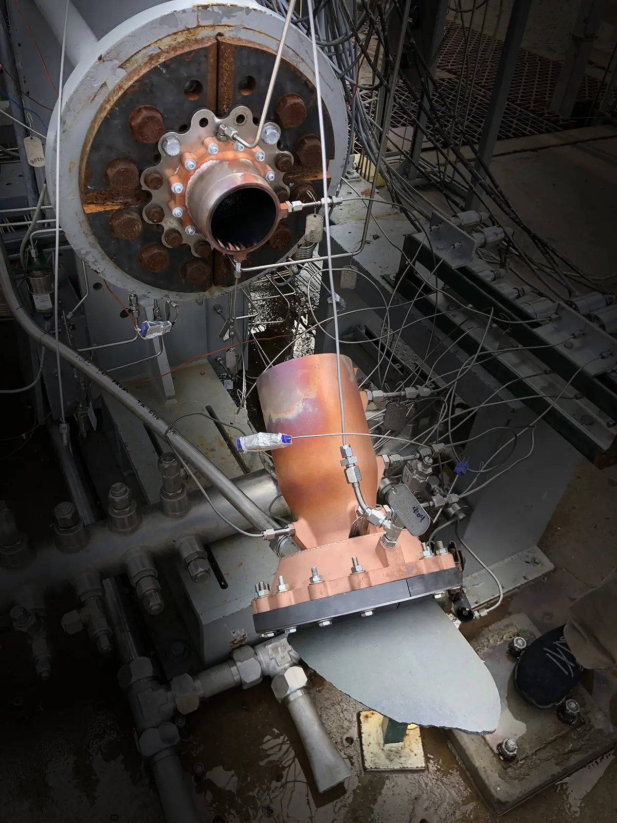The copper-colored 3D-printed combustion chamber on the test stand after the failed engine test, surrounded by instrumentation lines, with a broken grey section below its lower flange.