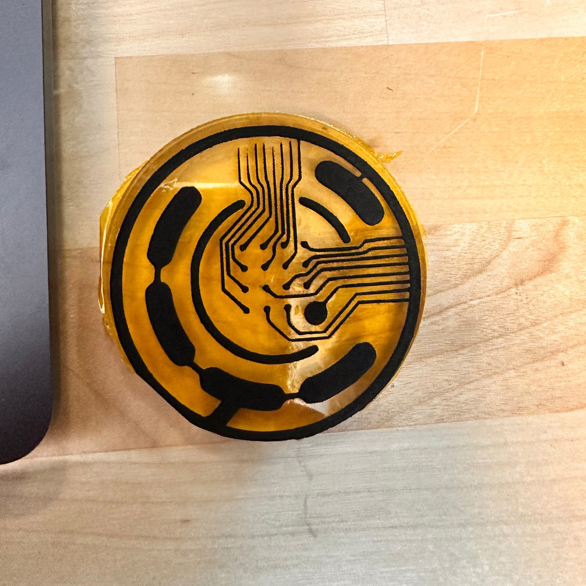 Circle of orange Kapton film with black laser-drawn lines: two bundles of thin wires ending in small dots near the center, inside a ring of wide curved shapes